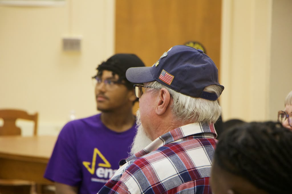 A member in a purple Enactus shirt and a veteran in a flag cap listening at a community meeting